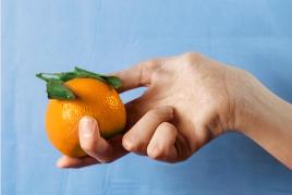 Michelle Miles, hand model, 2018, Filmstill. Bildbeschreibung: Eine Frauenhand hält eine Orange vor einem hellblauen Hintergrund