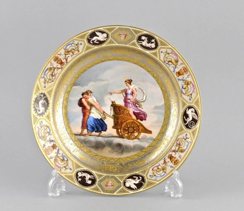 Biedermeier-Gemäldeteller, Wien, aus der Zeit zwischen 1810-1819, aufwendig dekoriert mit Fabelwesen und Maskaronen, zeigt im Spiegel die Jagdgöttin Diana auf einem von zwei jungen Frauen gezogenen Streitwagen. Das Gemälde 'Diana als Mondgöttin im Streitwagen' von Raffael diente hierfür als Vorlage.(Limit 3.500 €)